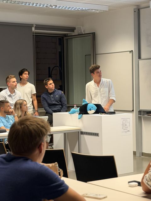 A member speaking at the front of a classroom while others watch, with a blue plush shark on the desk