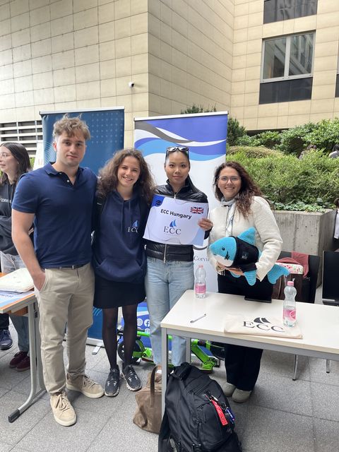 Four smiling people at the ECC Hungary stand in a university courtyard, one holding a sign and a plush shark